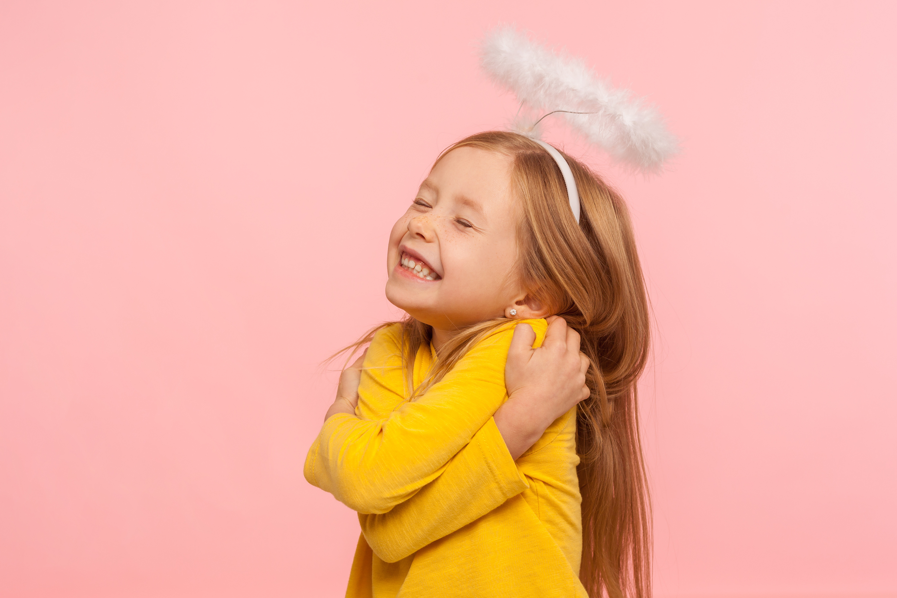 Giggling Girl with Halo Embracing Herself on Pink Background